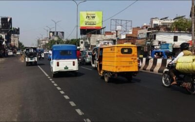 Billboard placed by hoarding advertising companies in Lucknow on a crowded commercial road with autos, bikes, and daily commuter traffic.